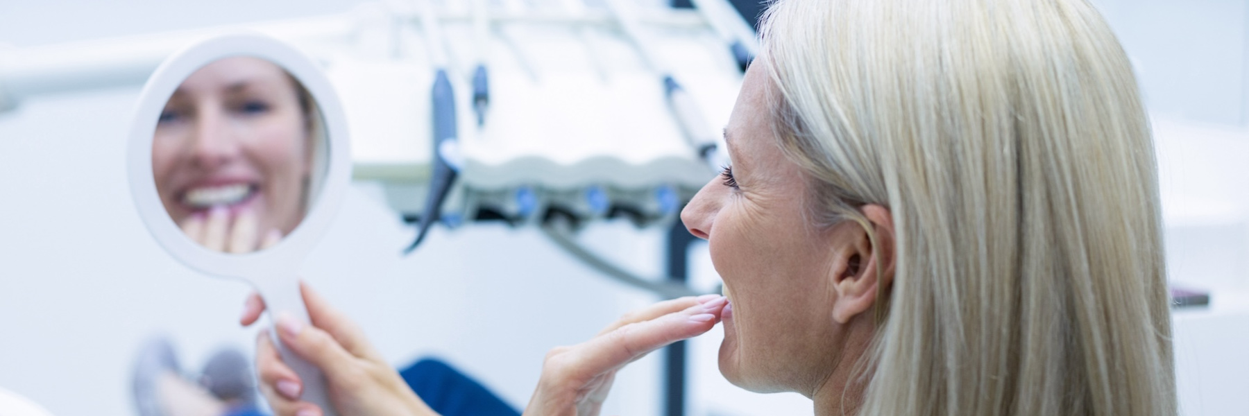 Older woman smiling while examining her teeth in a hand mirror at a dental office